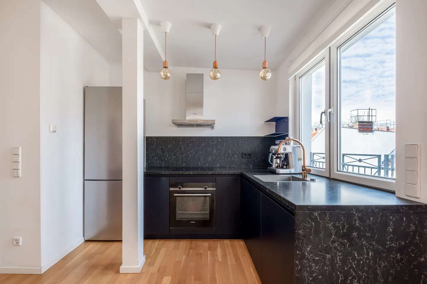 Bright kitchen with stainless steel fridge, dark cabinets, marble counters, and wood floors. Three pendant lights hang above.