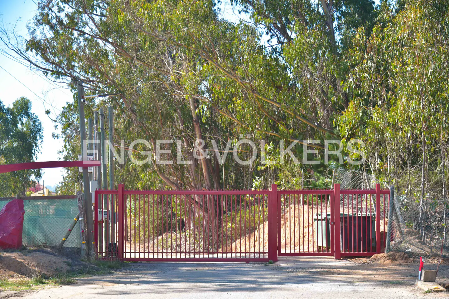 Red metal gate with Engel & Völkers logo in the background, surrounded by trees and greenery.