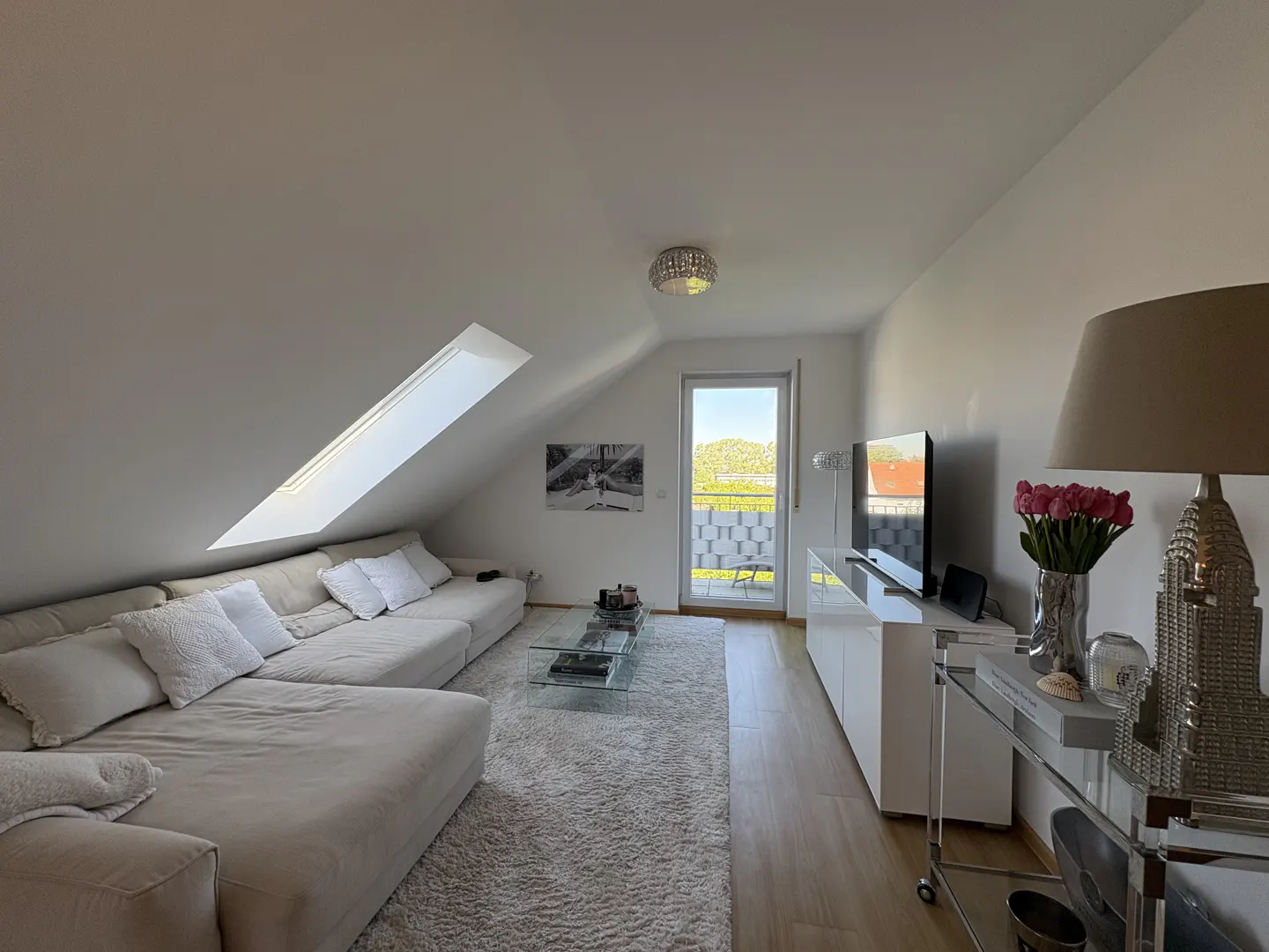Bright living room with a large white sectional sofa, glass coffee table, and a skylight. A balcony door offers a view of greenery.