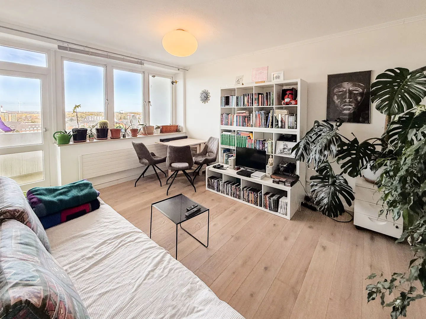 Bright living room with a sofa, black table, and a white bookshelf filled with books and a TV. Large windows offer a city view.