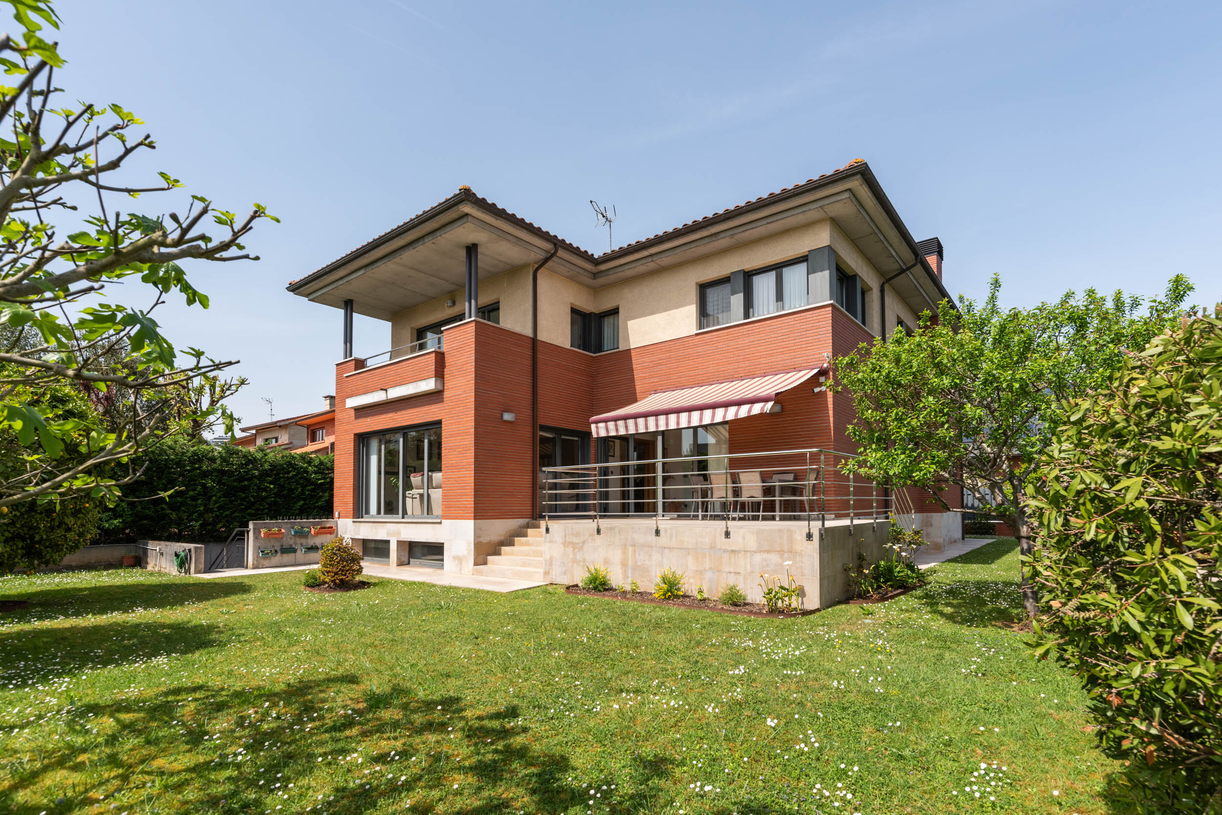 Two-story house with brick and beige siding, a balcony, and a patio with a red and white awning, surrounded by a green lawn and trees.