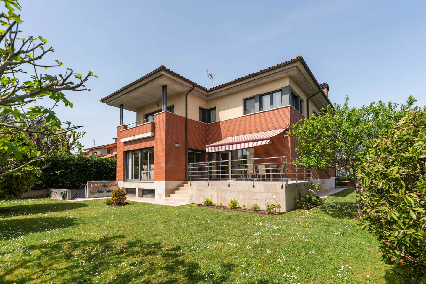 Two-story house with brick and beige siding, a balcony, and a patio with a red and white awning, surrounded by a green lawn and trees.
