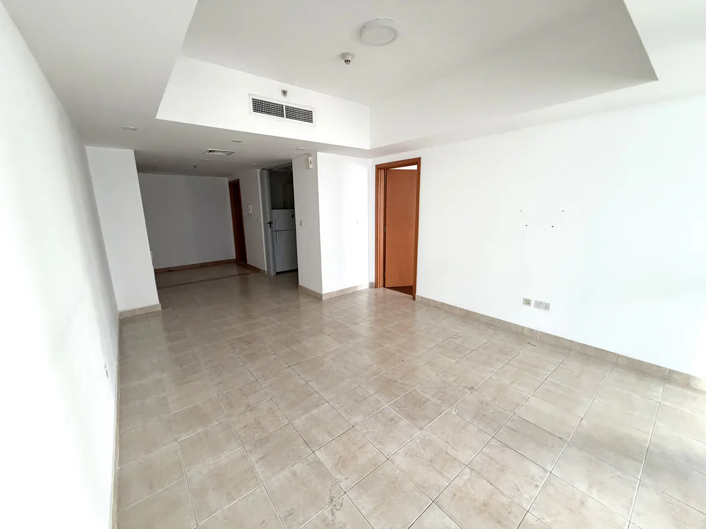 Empty room with white walls, beige tile floor, and a brown door. A glimpse of the kitchen with a white refrigerator is visible.