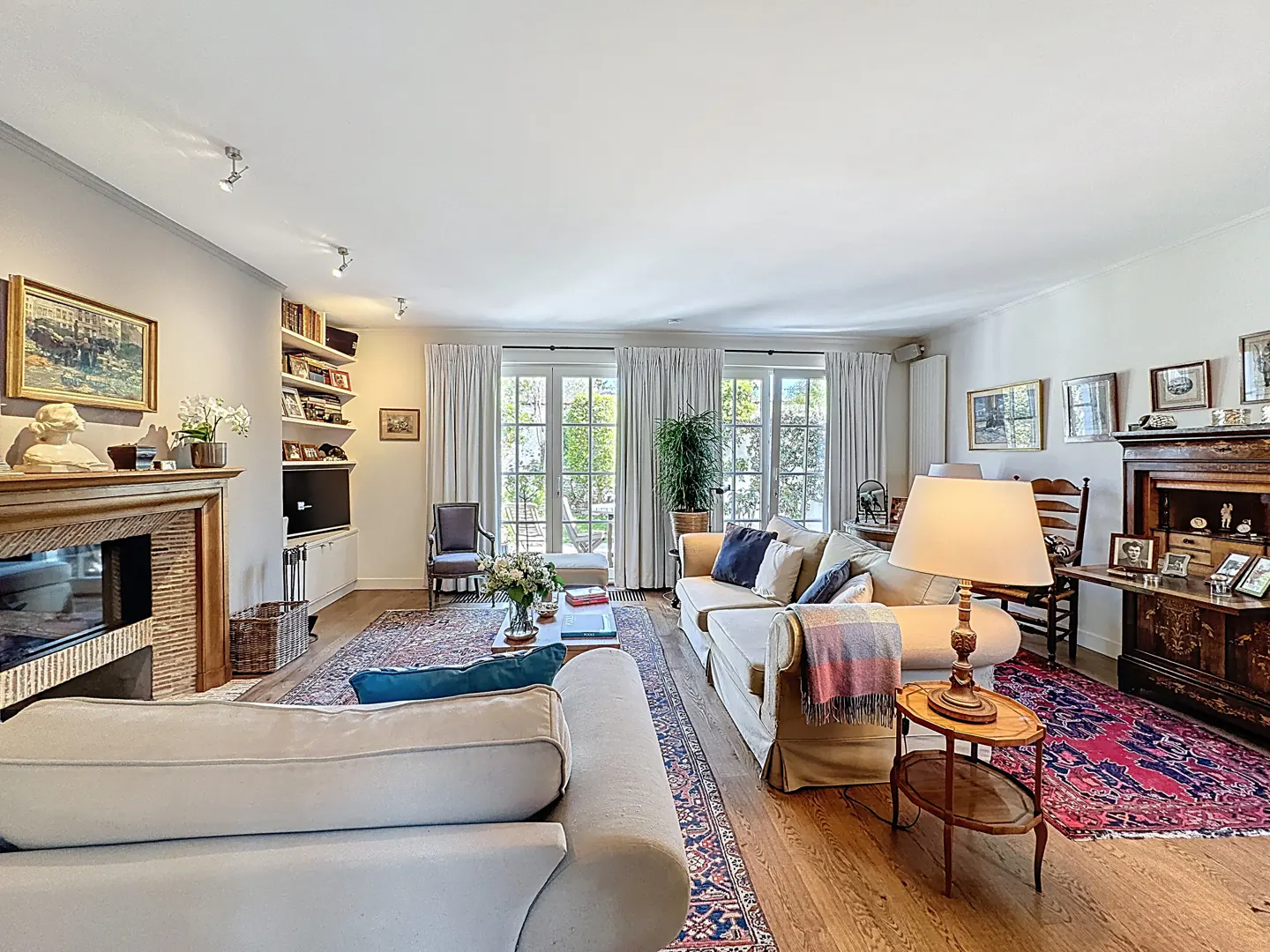 Bright living room with hardwood floors, white sofas, and French doors leading to a garden. A fireplace and built-in shelves add character.