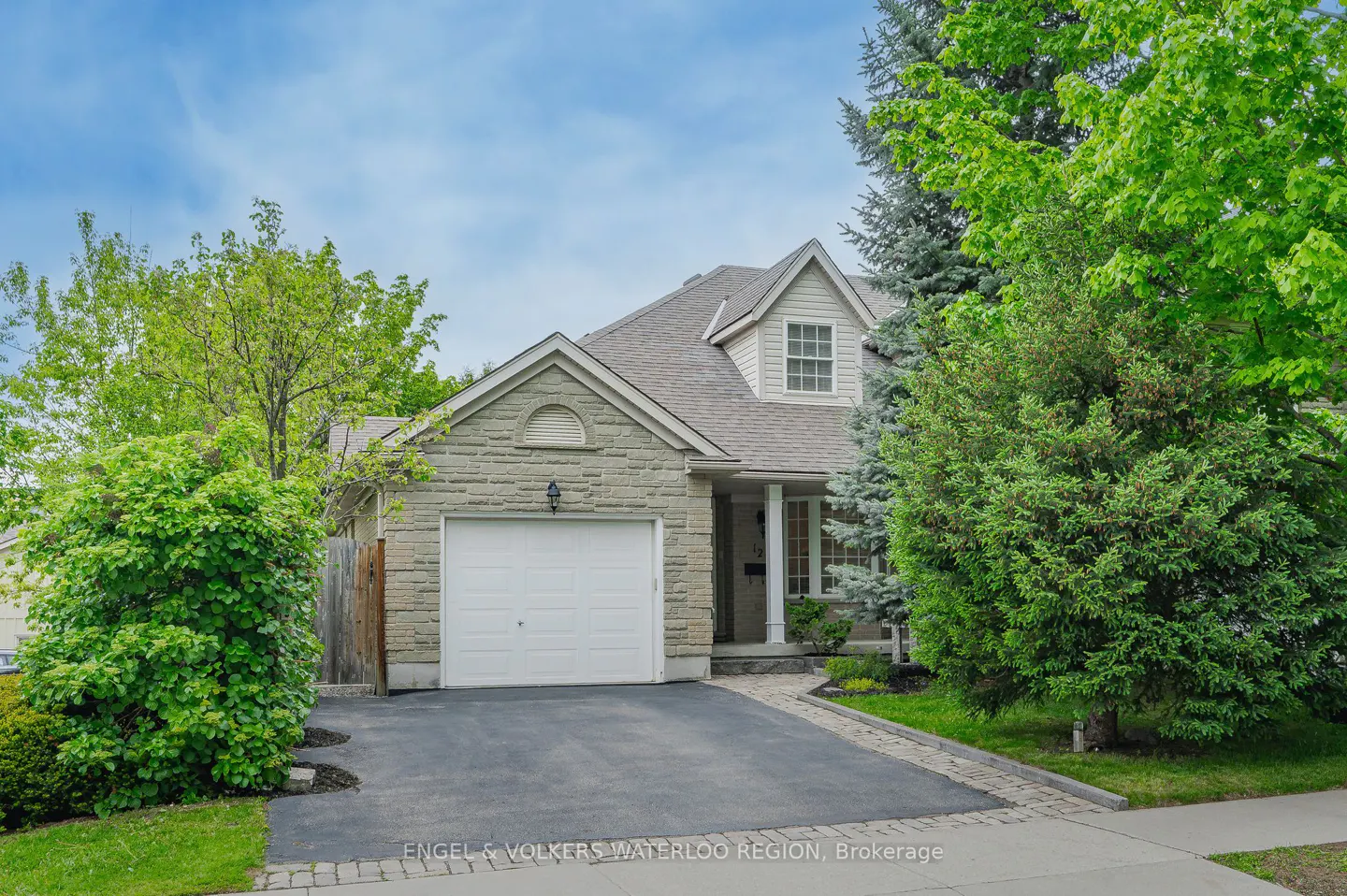 A one-story stone house with a white garage door and green trees under a blue sky.