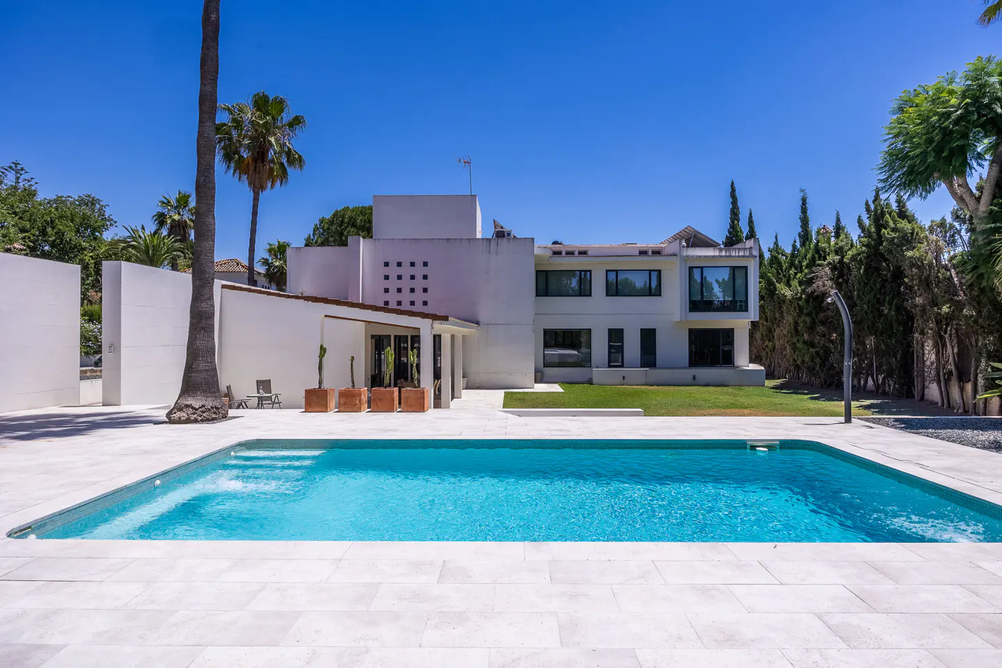 Modern white villa with a swimming pool, patio, palm trees, and green lawn under a clear blue sky.