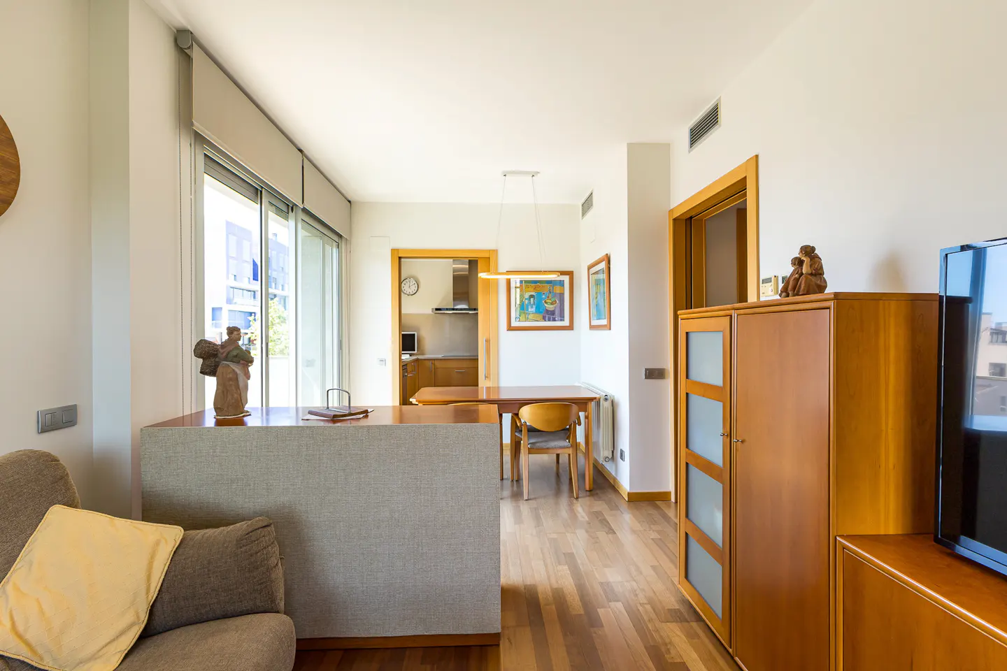 Bright apartment interior with wood floors, a gray sofa, and a wood dining table. A kitchen is visible in the background.