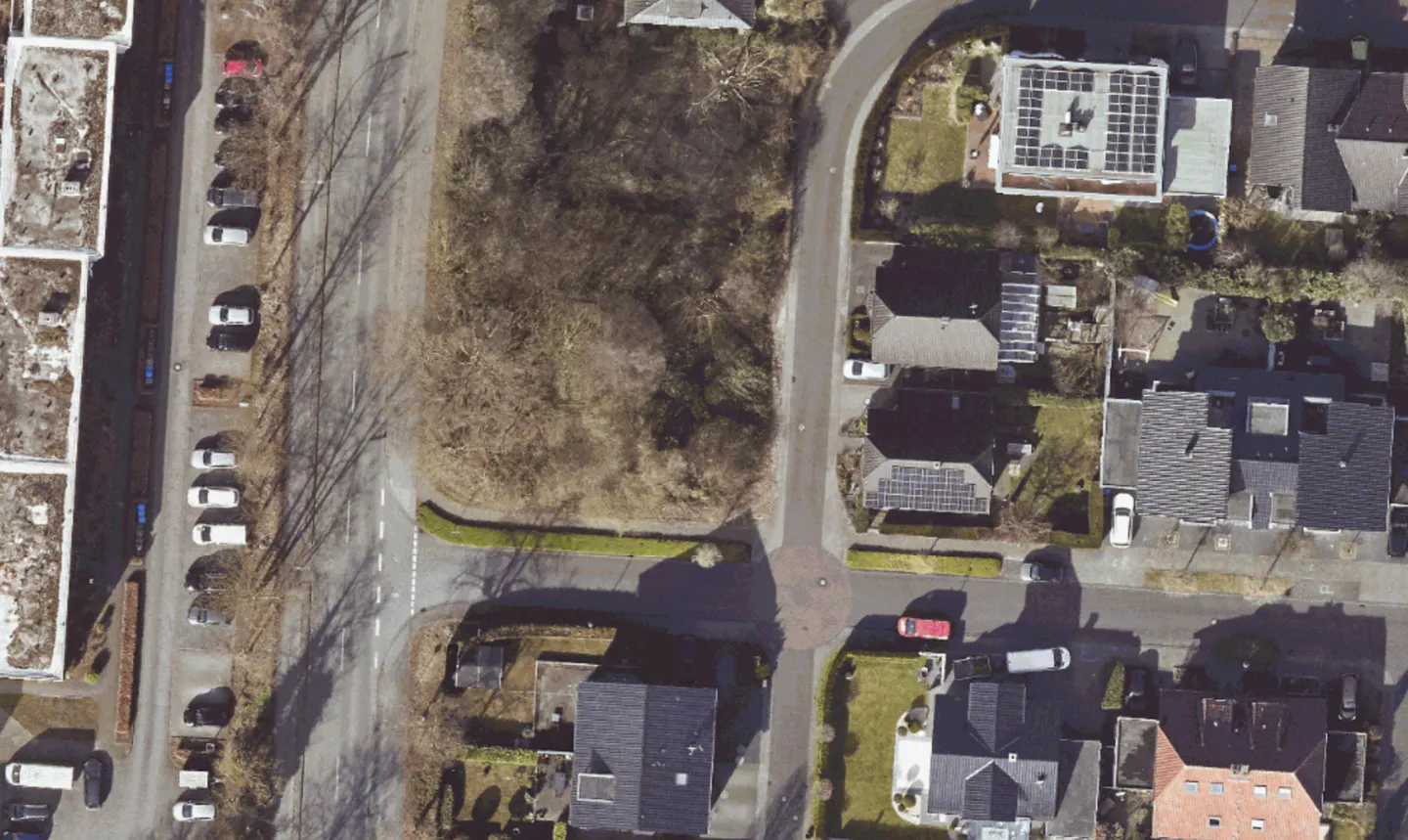 Aerial view of a vacant lot surrounded by roads, houses, and parked cars. Some houses have solar panels on their roofs.