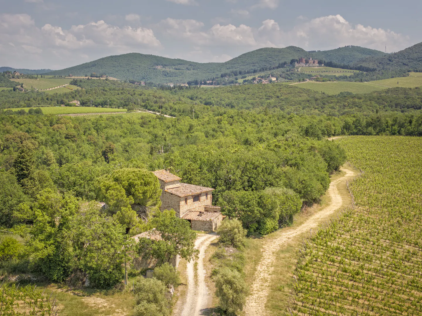 Aerial view of a stone farmhouse nestled in a lush Tuscan landscape with vineyards, dirt paths, and distant rolling hills.