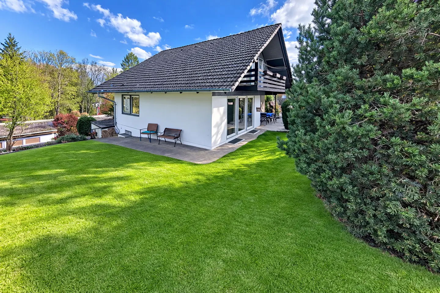 White house with a dark tiled roof, a patio with benches, and a large green lawn under a blue sky with scattered clouds.