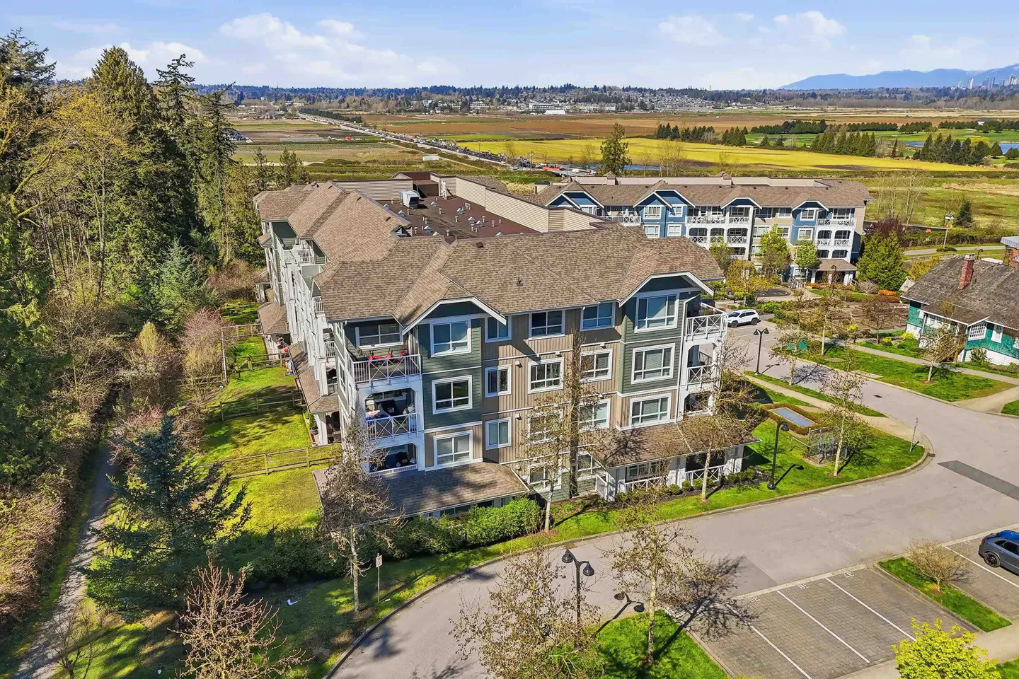Aerial view of a multi-story apartment building with green and tan siding, brown roof, and balconies, surrounded by trees and green space.
