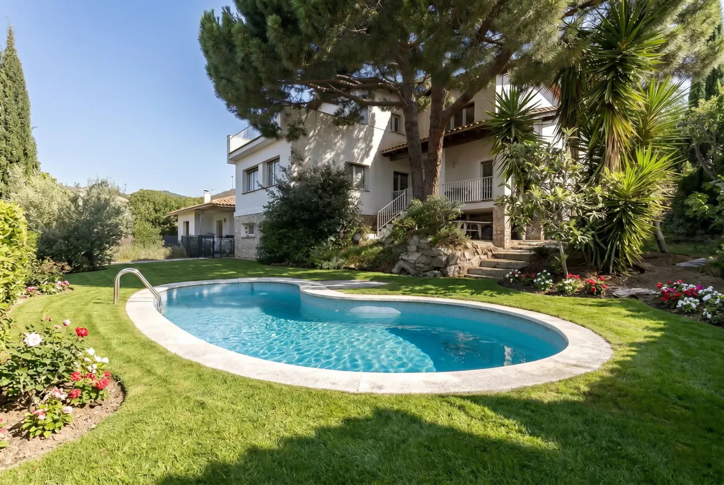 A kidney-shaped pool with turquoise water sits in a green lawn, with a white house and trees in the background.