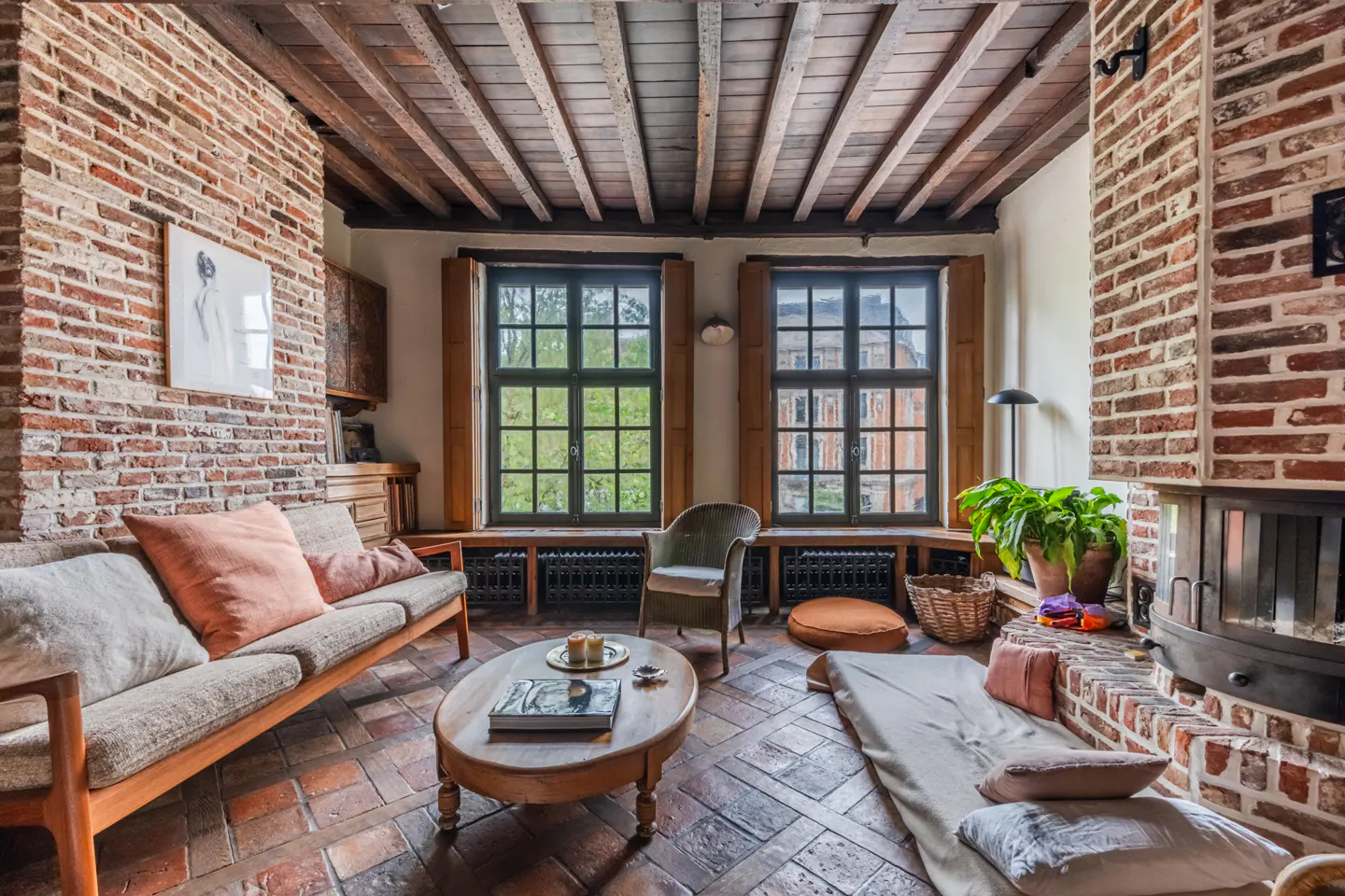 Cozy living room with exposed brick walls, wood-beamed ceiling, terracotta tile floor, fireplace, and two large windows.