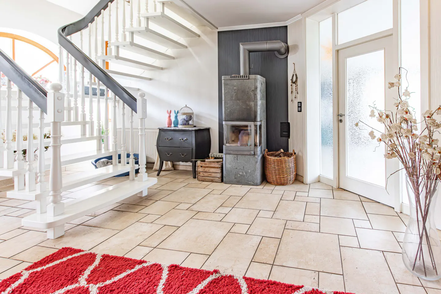 Bright entryway with white staircase, stone fireplace, tiled floor, red rug, and a glass vase with dried flowers.