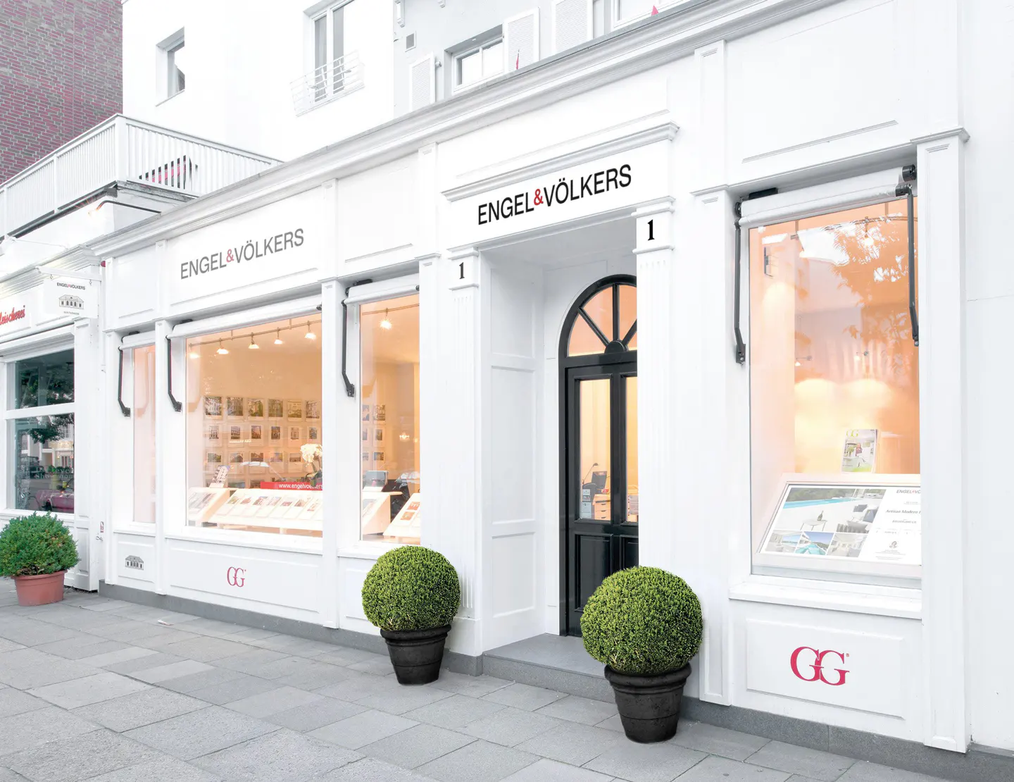 Exterior of Engel & Völkers real estate office with white facade, black door, and window displays. Topiary plants flank the entrance.