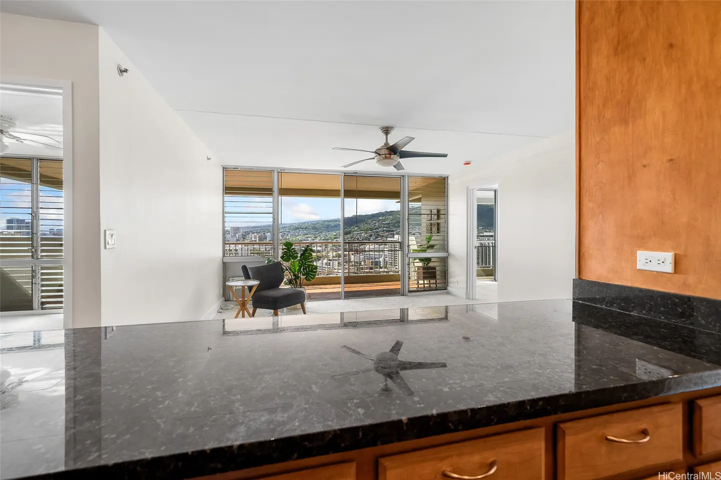 View from a kitchen counter with black granite looking toward a living room with a gray chair, ceiling fan, and city view.