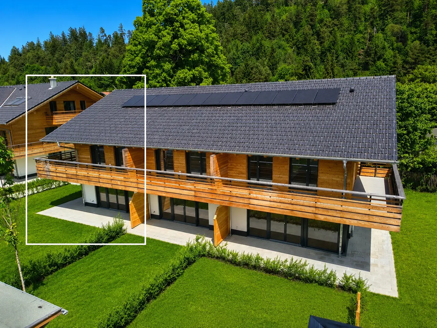 Aerial view of a modern wooden house with a dark tiled roof, solar panels, and a balcony, set against a lush green forest.