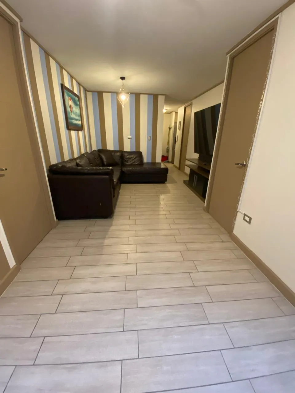 Living room with brown leather sectional sofa, striped walls, and light wood-look tile flooring. A TV and console are visible in the background.