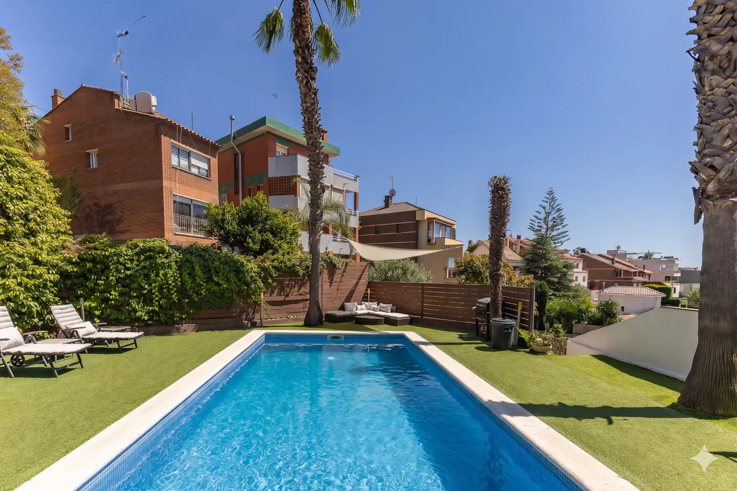 A backyard with a blue tiled pool, green lawn, lounge chairs, and palm trees, with houses in the background.