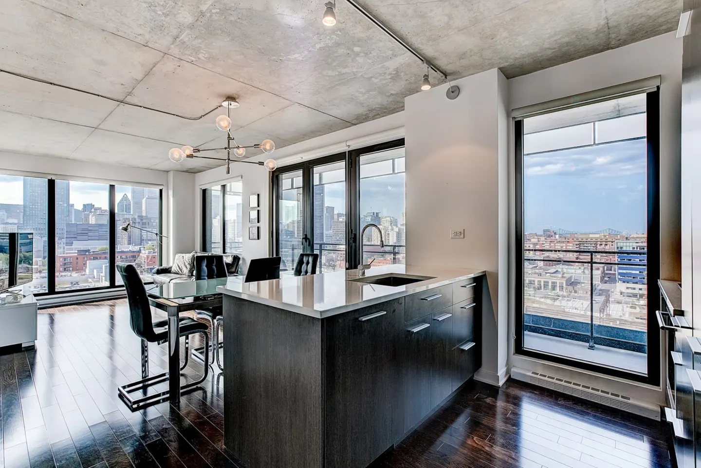 Modern condo interior with dark wood floors, a kitchen island, and floor-to-ceiling windows offering a city view.