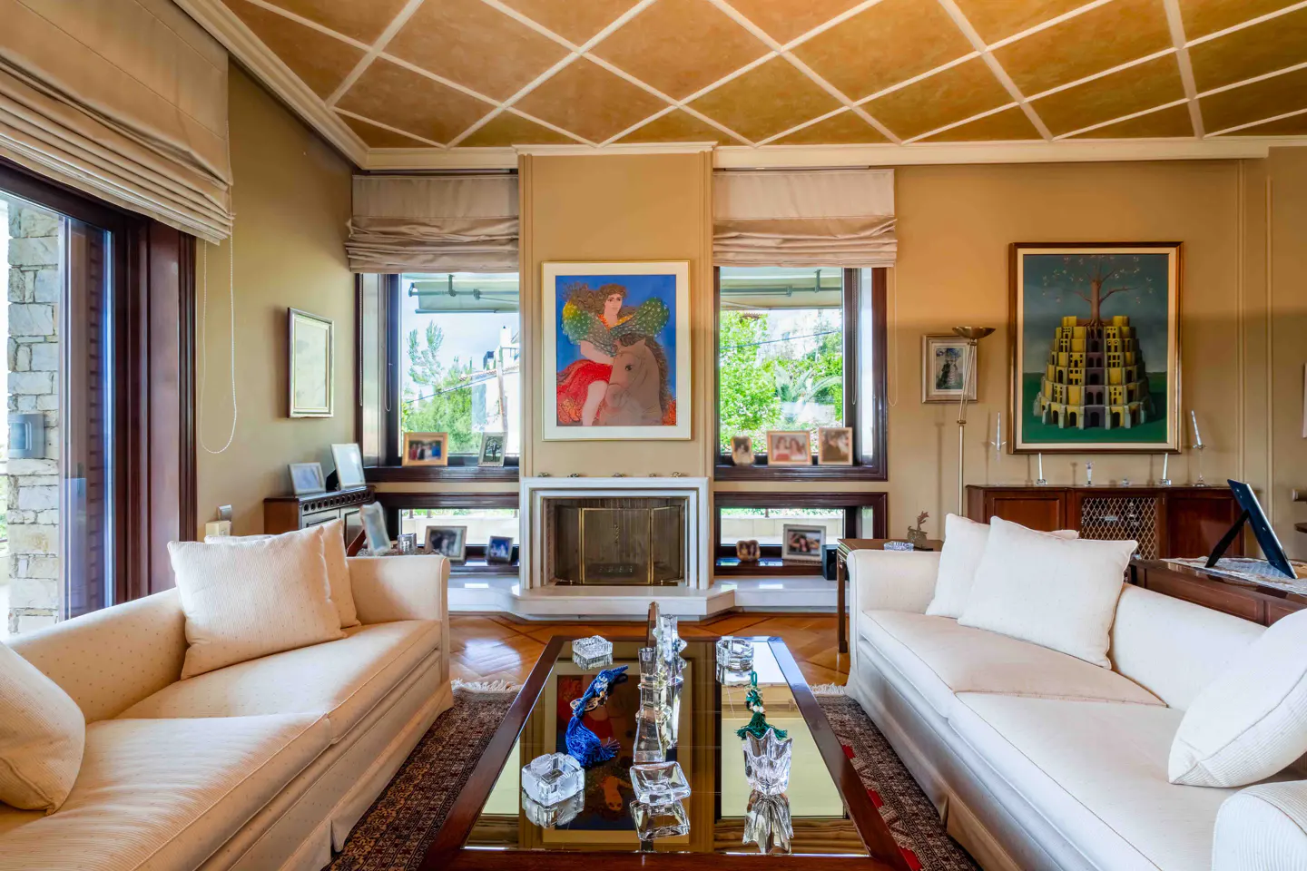 Living room with two white sofas, a glass coffee table, and a fireplace. Artwork hangs above the fireplace and on the wall. Windows with blinds let in natural light.