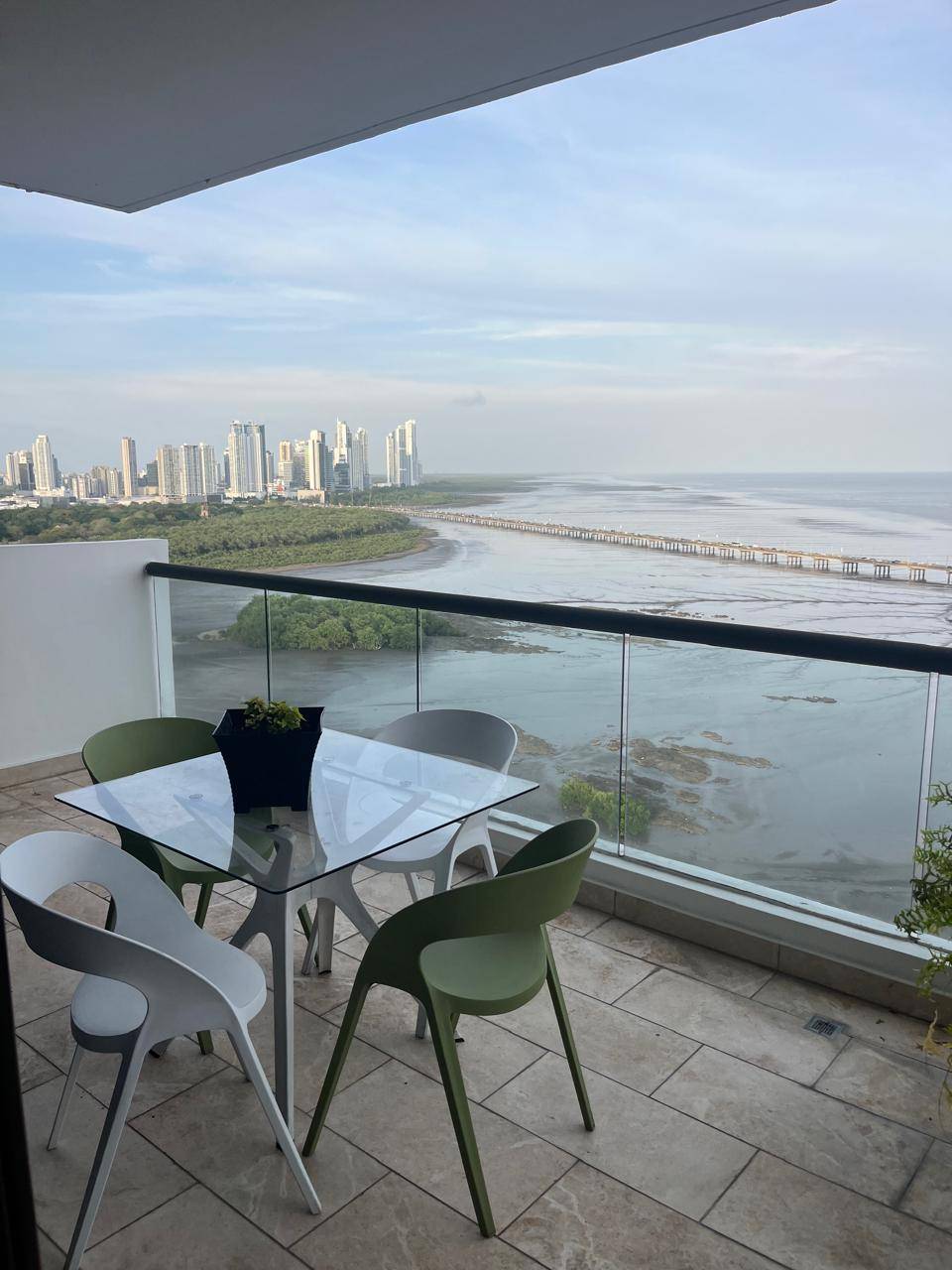 Balcony view with glass table, green and white chairs, overlooking ocean and city skyline.