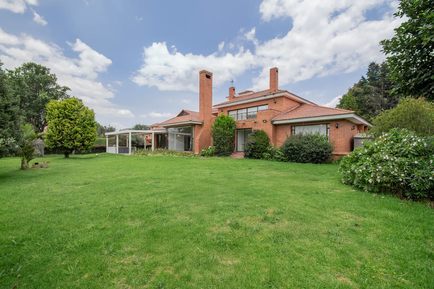 Brick house with a red tile roof, tall chimneys, and a large green lawn under a cloudy sky.