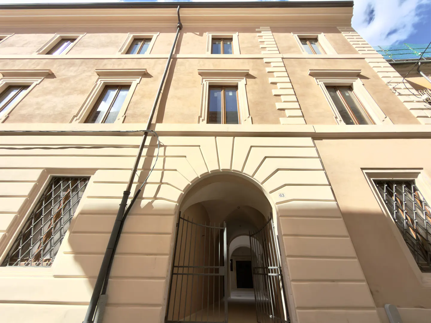 Exterior view of a beige building with arched entrance and metal gate. Windows with decorative trim and metal bars are visible. A drainpipe runs along the facade.