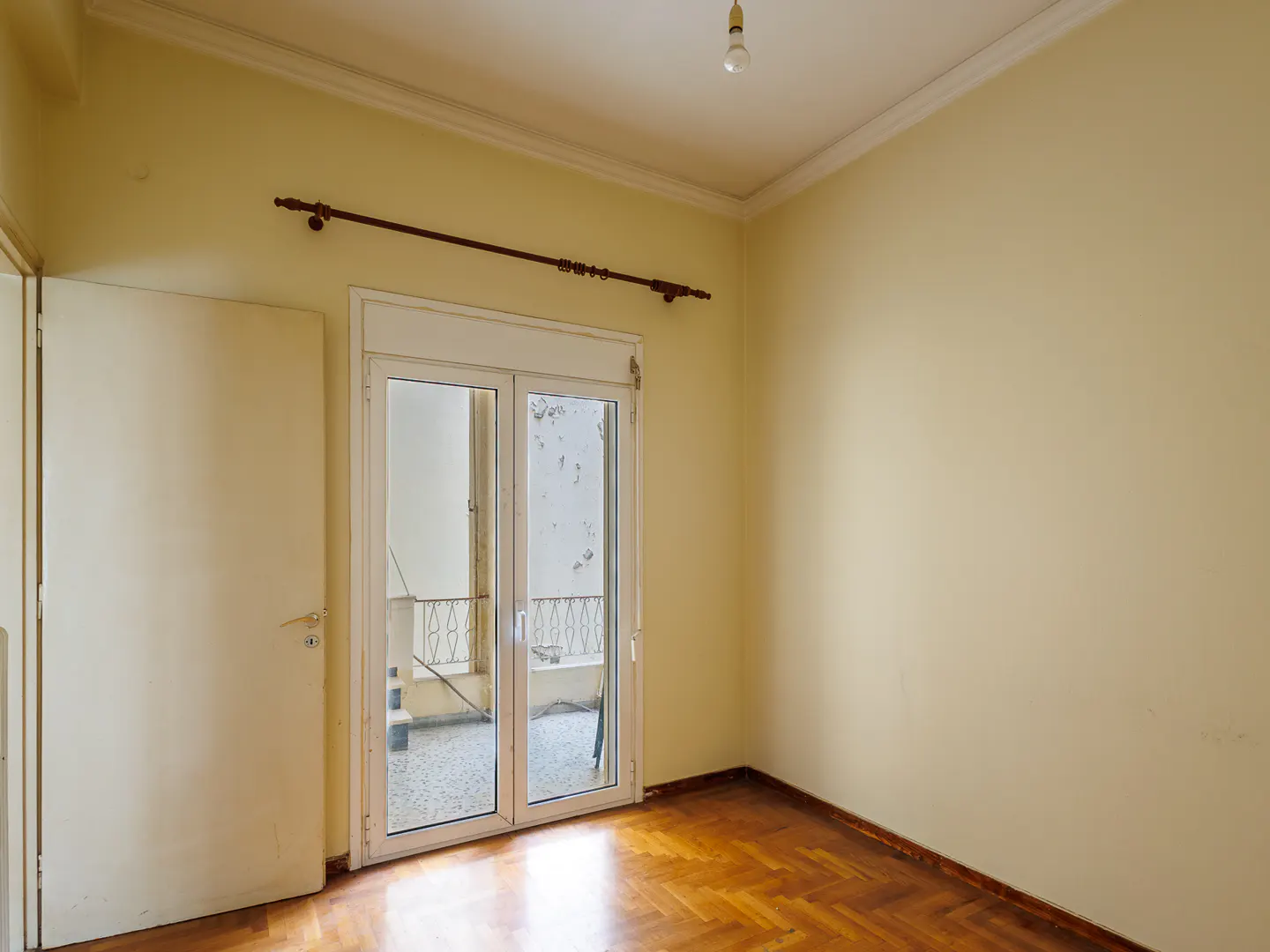 Empty room with cream walls, herringbone wood floors, a white door, and glass balcony doors with a curtain rod above.