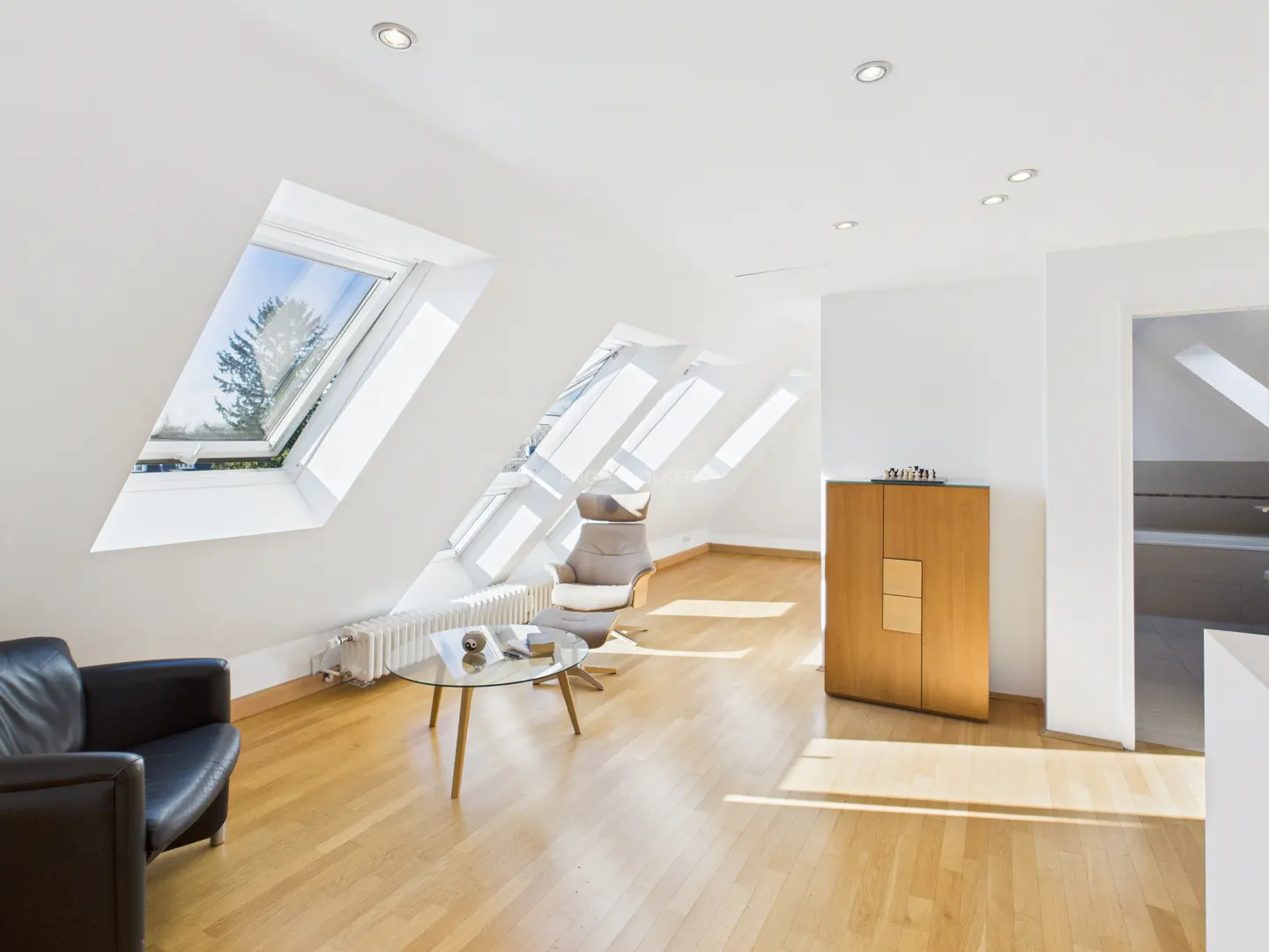 Bright attic living room with hardwood floors, white walls, skylights, black chair, and light wood cabinet.