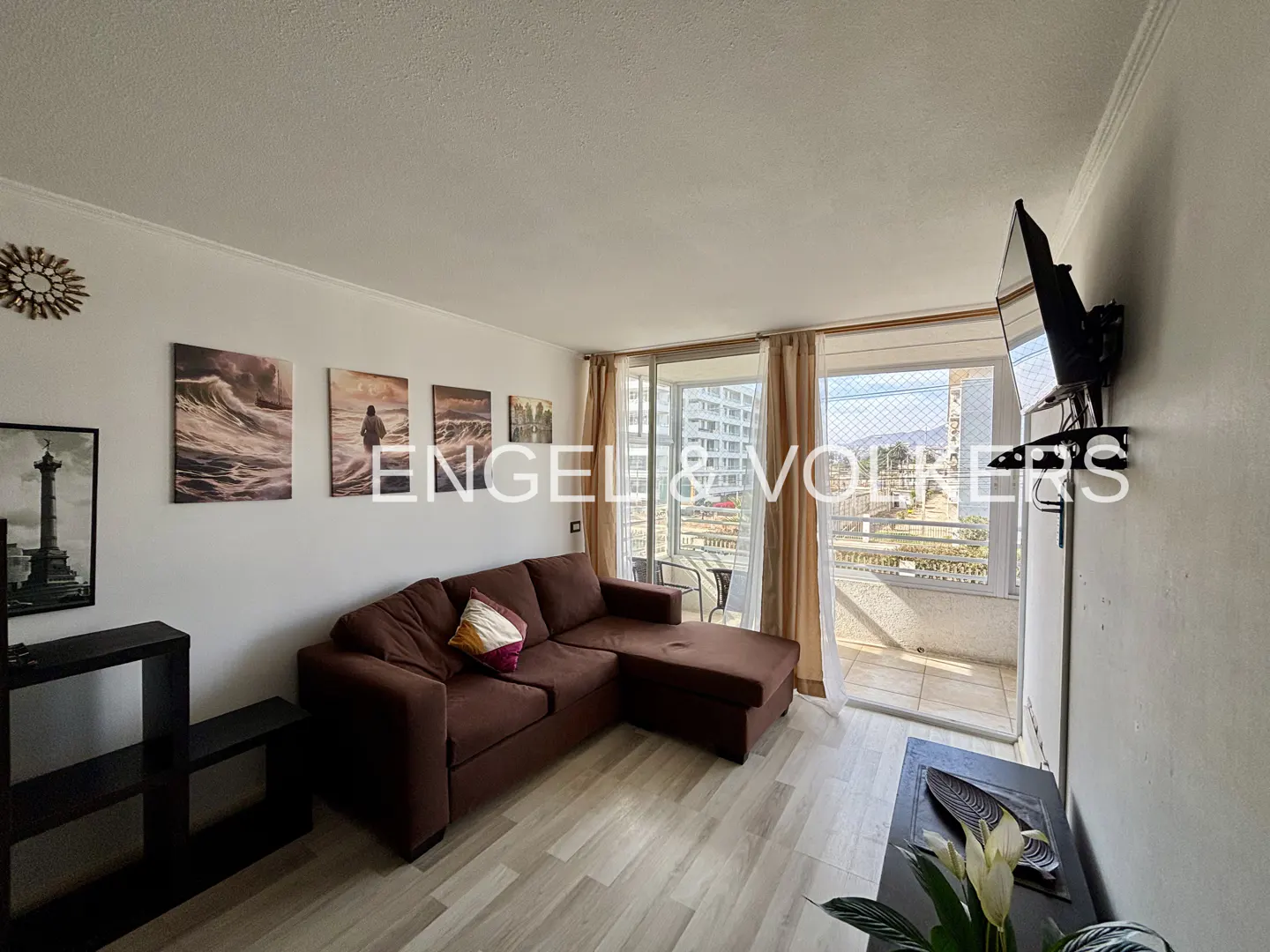 Living room with brown sectional sofa, wall art, and balcony with city view. TV mounted on the wall.