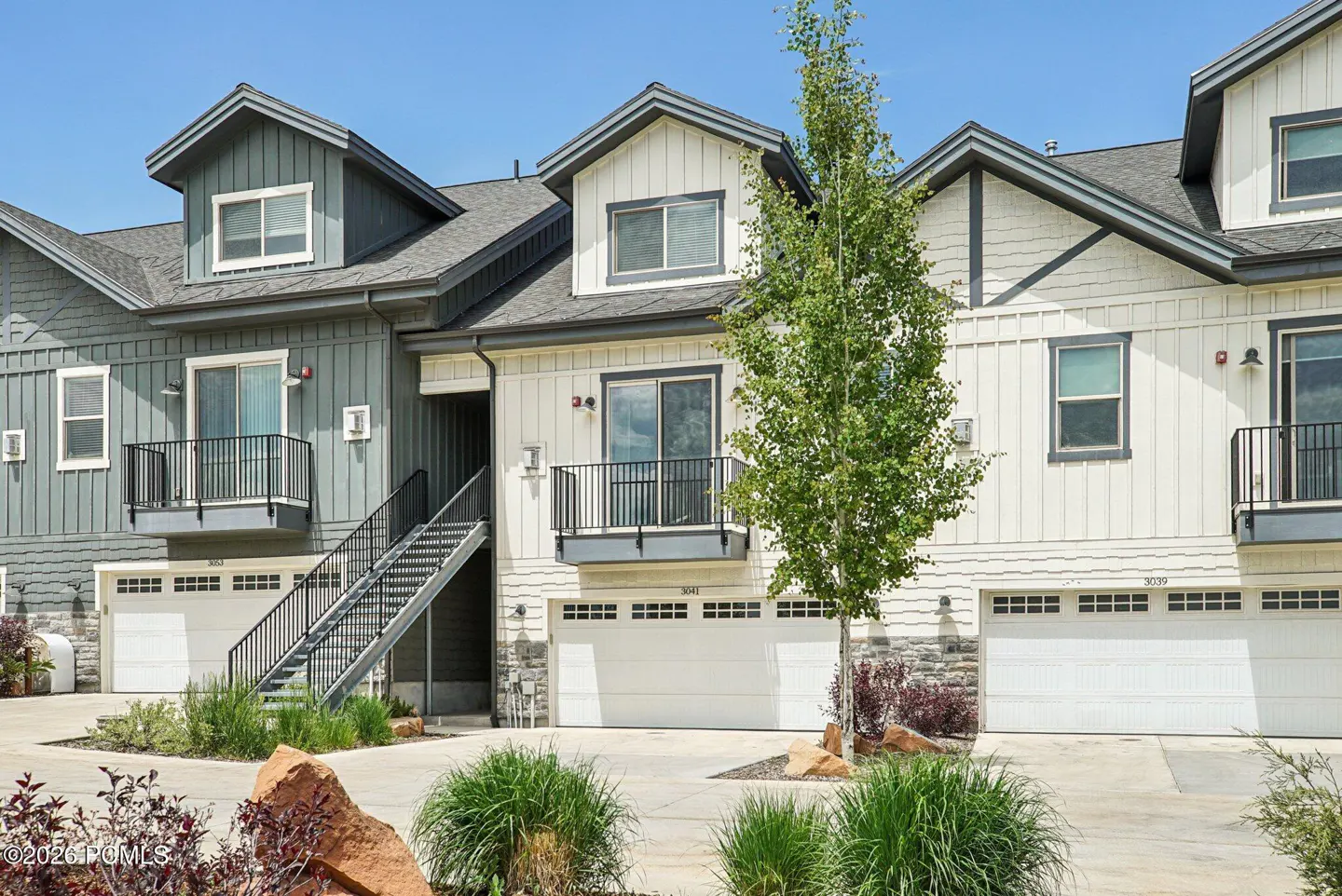 Exterior view of townhouses with white garage doors, gray trim, and black railings. A tree is in front of the middle unit.