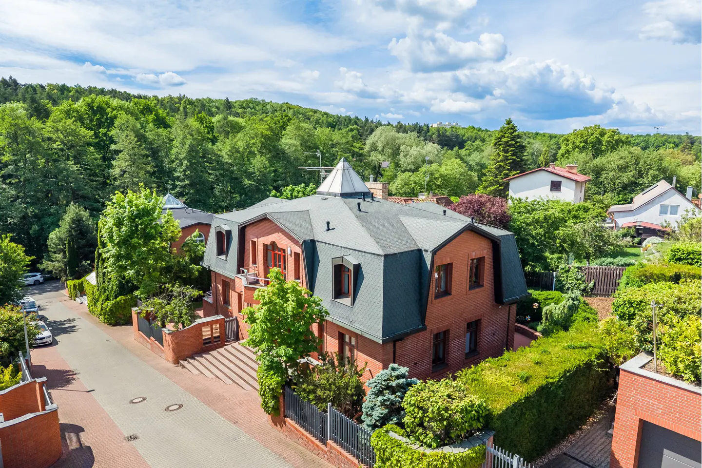 Aerial view of a red brick house with a dark roof and pyramid-shaped skylight, surrounded by lush trees and a quiet street.