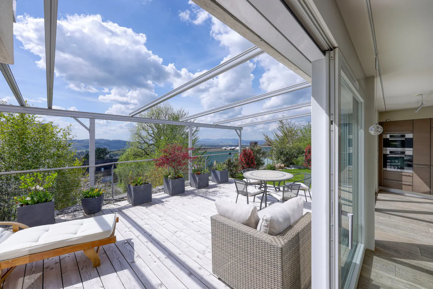 Bright, airy patio with white wood flooring, lounge chair, and dining set. Green trees and blue sky visible beyond the railing. Modern kitchen inside.