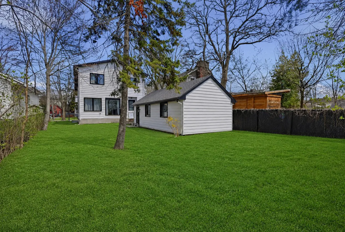 Backyard view of a white house and shed with green lawn, black fence, and trees under a blue sky.
