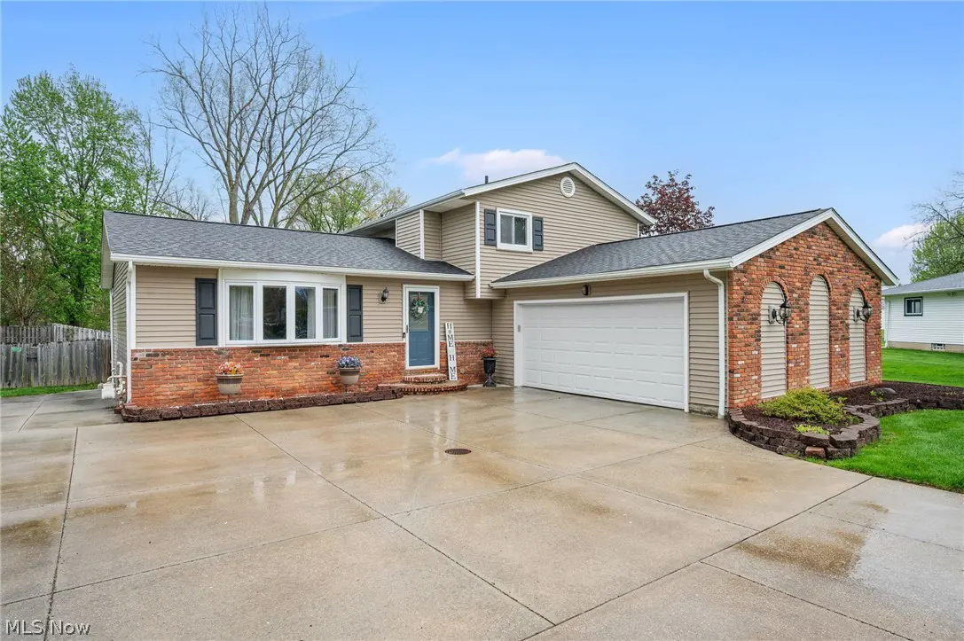 Two-story house with tan siding, brick accents, and a gray roof. A white garage door is on the right. The driveway is wet.