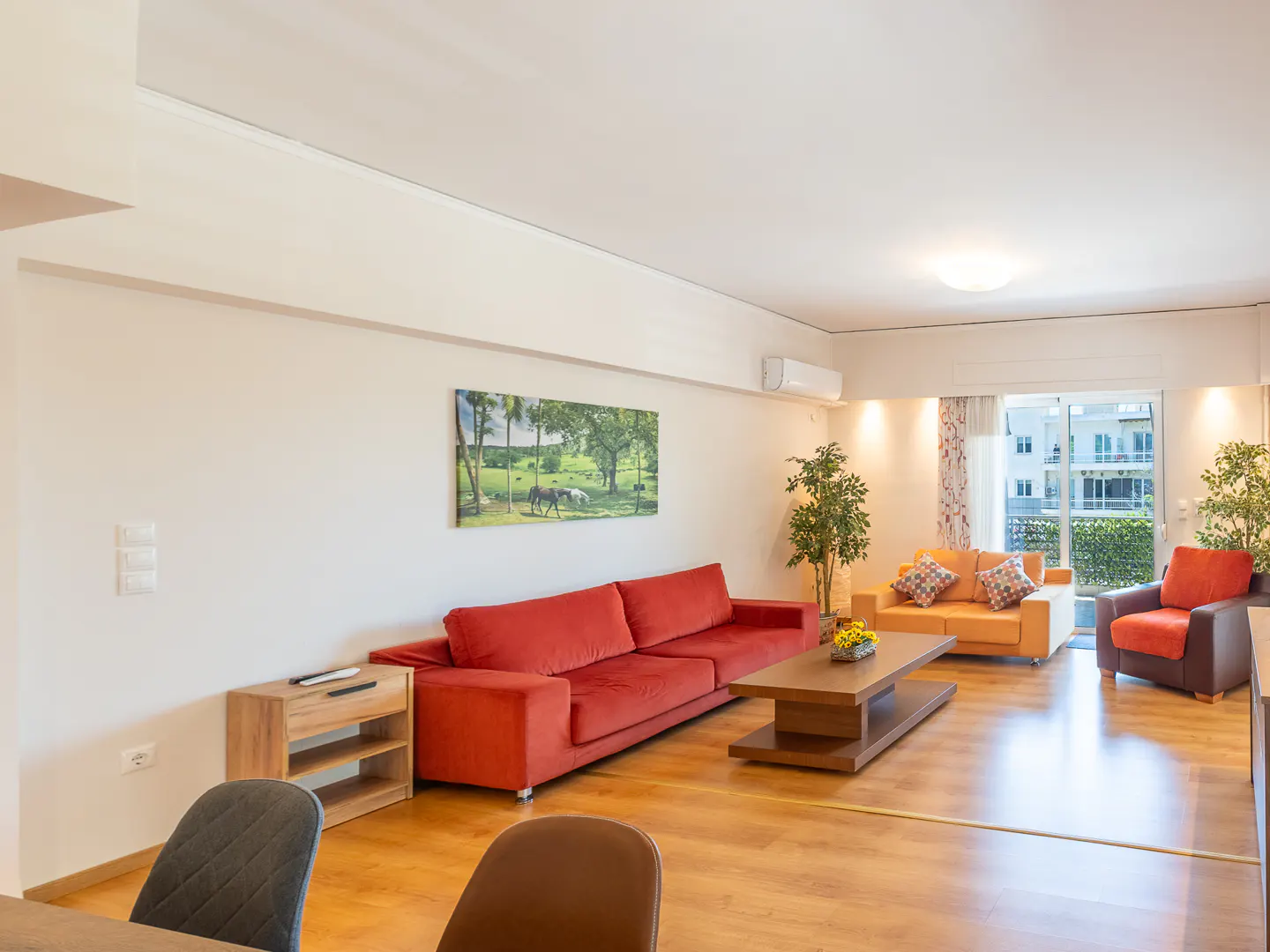Bright living room with a red sofa, orange loveseat, brown armchair, wooden coffee table, and a nature print on the wall.