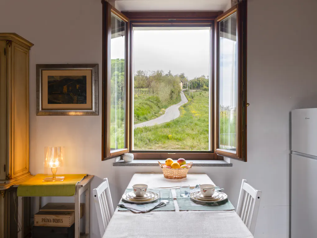 Cozy dining area with open window view. Table set for two, fruit basket, and a scenic landscape outside.