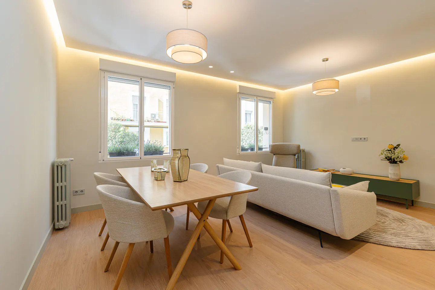 Modern living room with light wood floors, a dining table with four chairs, and a beige sofa under recessed lighting.