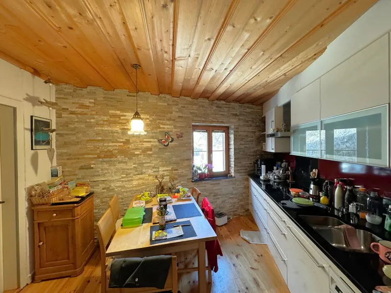 A kitchen with wood floors, a wood ceiling, a stone wall, and white cabinets. A table and chairs are in the center.