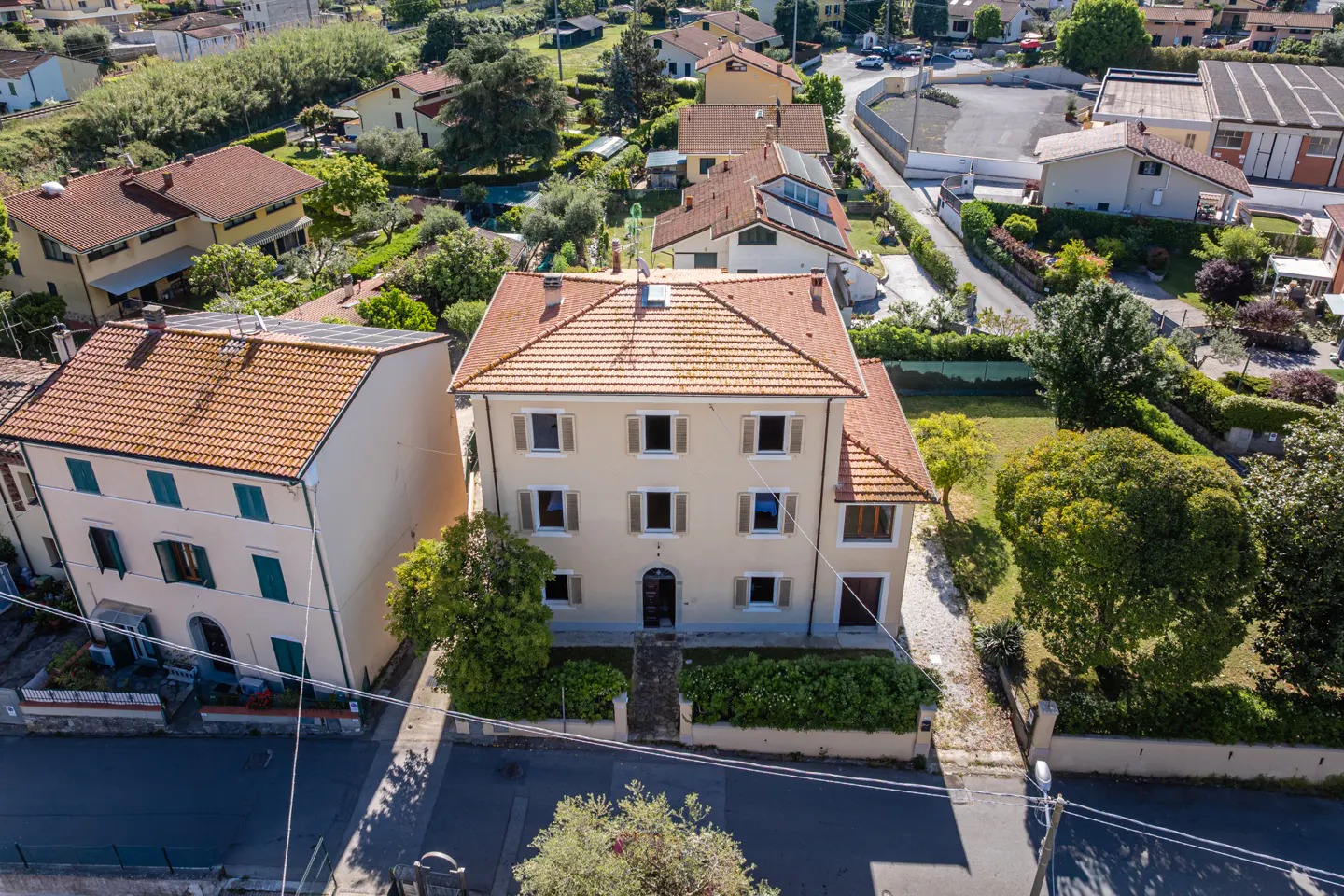 Aerial view of a three-story beige house with a red tile roof, surrounded by green trees and other houses in a neighborhood.