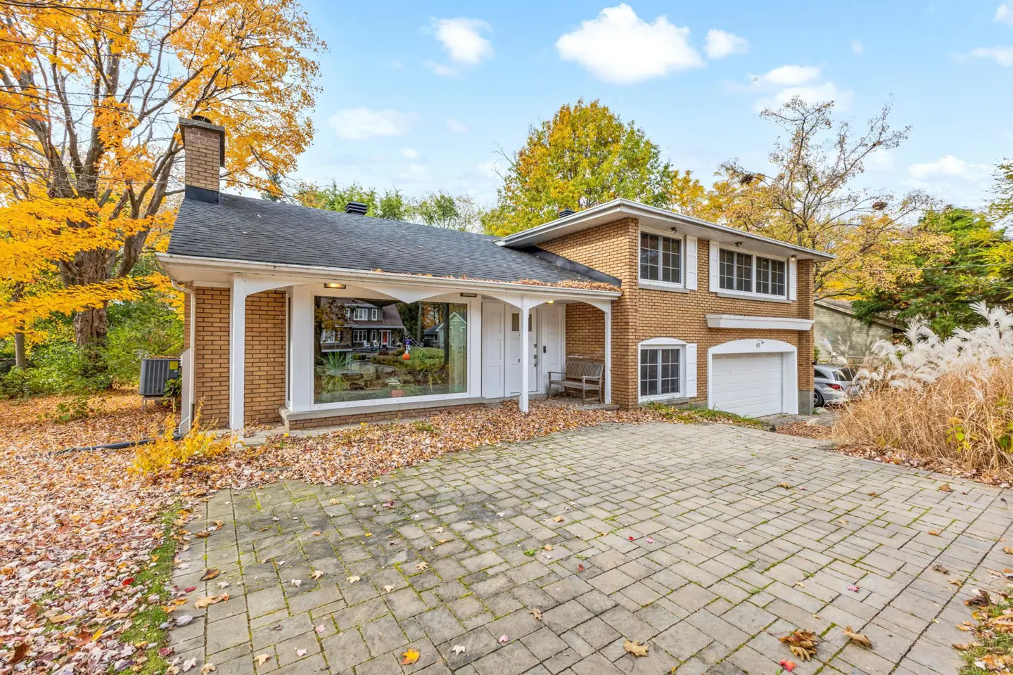 Two-story brick house with a black roof and white trim on a sunny autumn day. A stone driveway leads to the garage.
