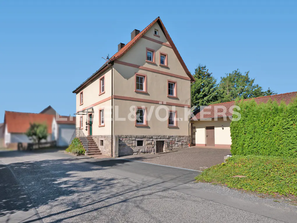 Two-story beige house with a red roof and trim, stone foundation, and a garage on a sunny day.