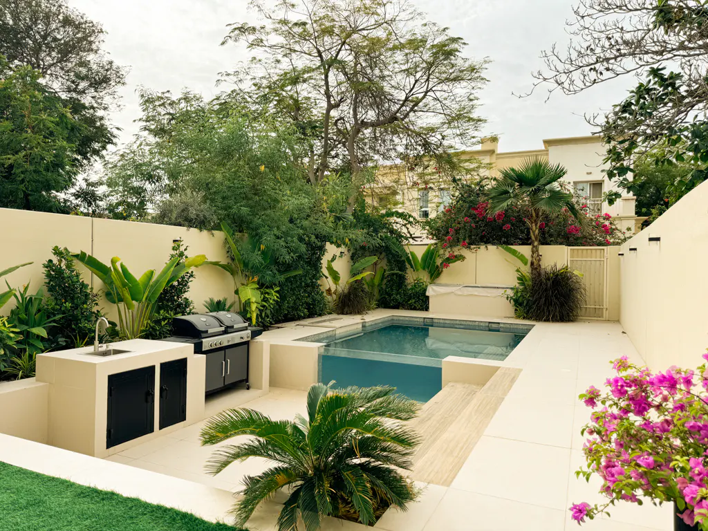 Backyard with a pool, outdoor kitchen with a grill, and lush greenery against a beige wall.