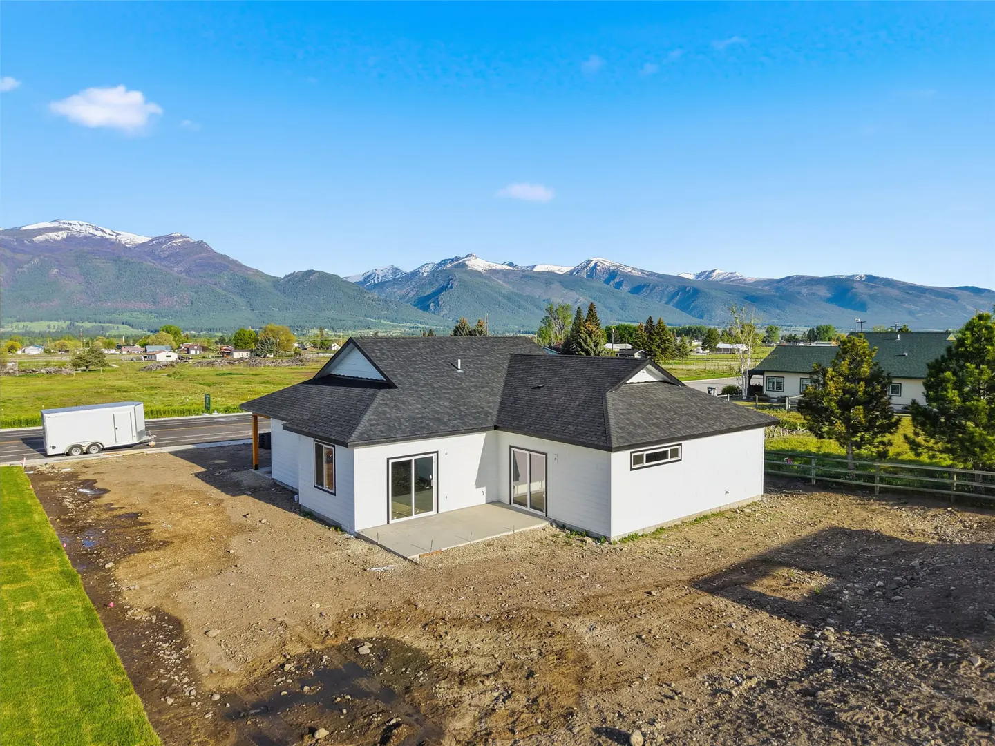 Aerial view of a modern white house with a dark roof, set against a backdrop of mountains and a blue sky.