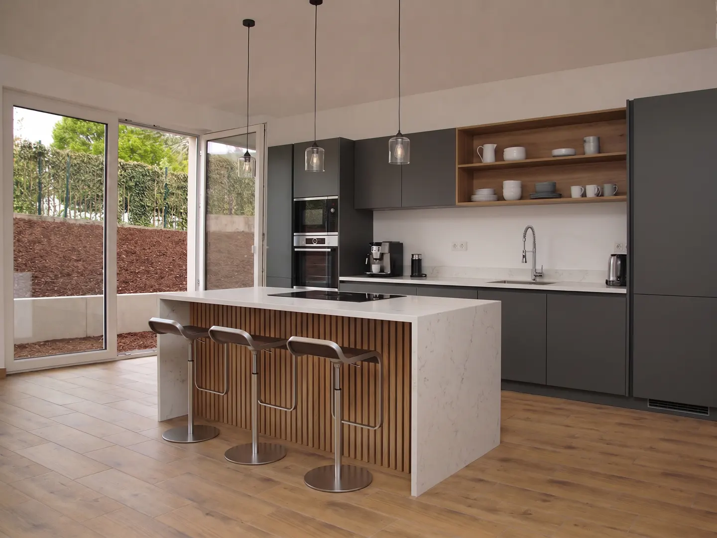 Modern kitchen with gray cabinets, white countertops, and a wood-paneled island with three stools. Large windows offer a view of the outdoors.
