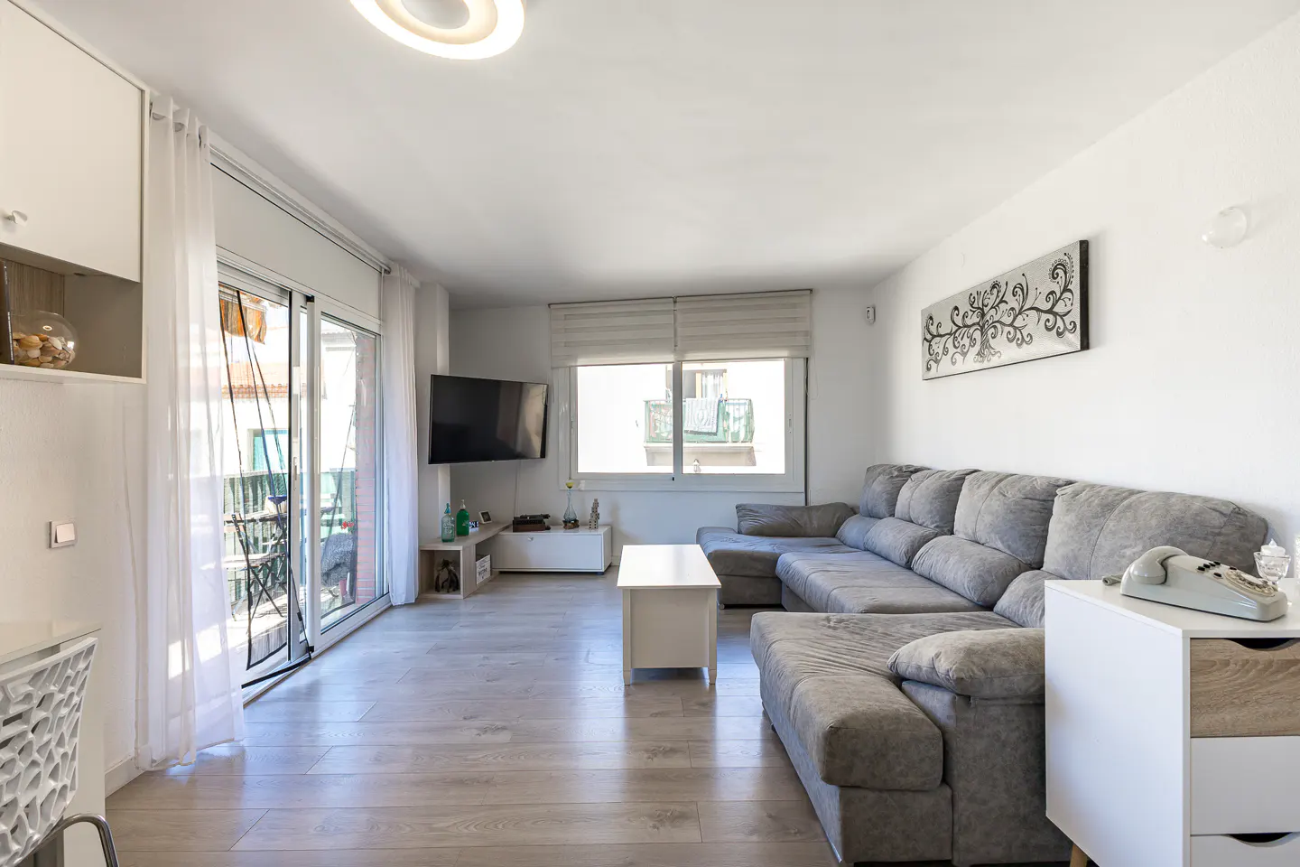 Bright living room with a large grey sectional sofa, white coffee table, wall-mounted TV, and balcony access.