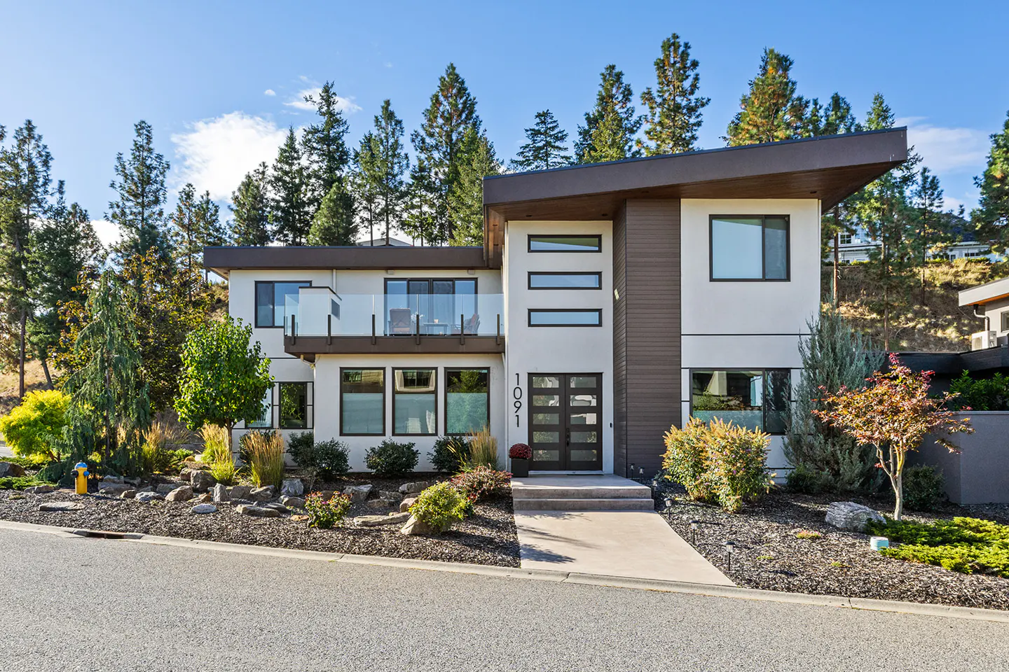 Modern two-story home with a balcony, white and brown exterior, black framed windows, and a landscaped yard.