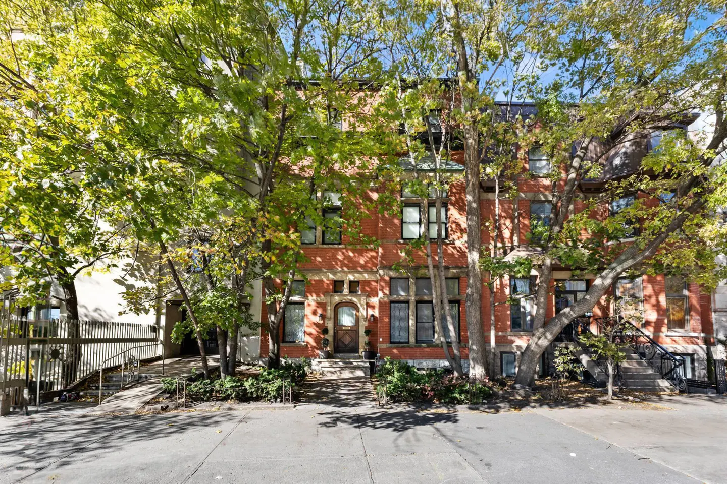 A three-story red brick building with black windows is partially obscured by green trees. A sidewalk is in the foreground.
