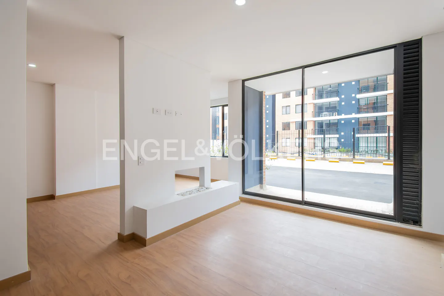 Bright, empty room with wood floors, white walls, and a modern fireplace. Large windows offer a view of a blue apartment building.